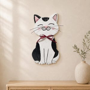 May include: A wall-mounted cat decoration featuring a white and black color scheme with a pink nose, pink-tinted cheeks, and a dark red ribbon tied around its neck. The cat has a smiling expression with closed eyes and thin wire whiskers.