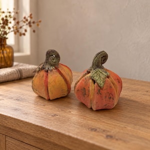 Pode incluir: Duas abóboras decorativas com um acabamento texturizado em laranja e amarelo. Cada abóbora apresenta um caule verde proeminente e curvado, com um design de corpo segmentado. As abóboras são arredondadas com cristas visíveis.