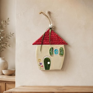 Puede incluir: Un adorno de pared de cerámica con forma de casa, techo rojo de escamas, cuerpo color crema, puerta arqueada verde oscuro con pomo blanco y una pequeña ventana azul con contraventanas verdes. Tres pequeñas flores pintadas decoran la parte inferior.