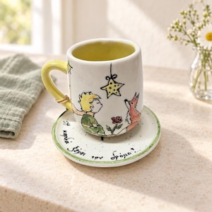 Könnte beinhalten: Eine weiße Keramiktasse mit passender Untertasse, verziert mit einer handgemalten Illustration eines kleinen Jungen mit gelben Haaren, einem Fuchs, einer Blume und einem hängenden Stern. Der Henkel ist gelb und die Untertasse zeigt griechischen Text.