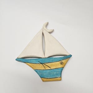 Può includere: Un ornamento da parete in ceramica a forma di barca a vela con due vele triangolari bianche e uno scafo decorato con strisce orizzontali in tonalità alternate di blu e giallo. Una piccola luna crescente sovrasta le vele e un timbro è visibile.