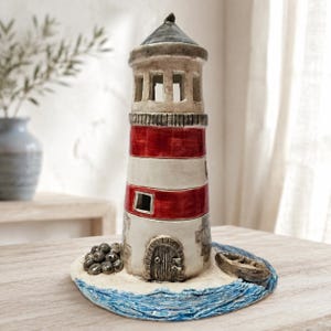 May include: A ceramic lighthouse sculpture featuring horizontal red and white stripes, a dark grey conical roof, and an arched door. The base depicts a sandy shore with a small boat and a cluster of rocks, surrounded by textured blue water.