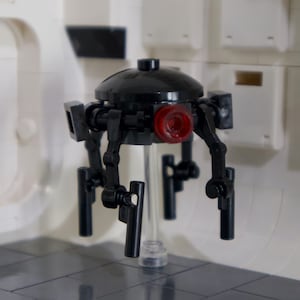 May include: A black and red toy droid with four legs and a round head. The droid is made of plastic and has a red light on the front.