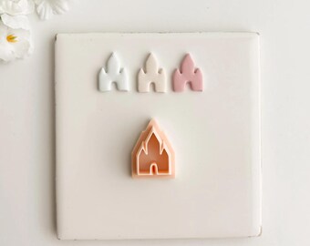 May include: A collection of castle-shaped cookie cutters in white, light pink, and pink hues. The cutters are arranged on a white square tile, with a sprig of white flowers in the background. These are ideal for creating themed cookies.