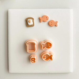 May include: A collection of food-themed cookie cutters in various shapes, including a slice of toast with an egg, a slice of bread, a flower, and fish. The cutters are a light orange color and are arranged on a white surface.