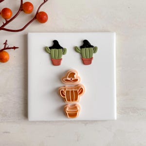 May include: A collection of cactus-shaped cookie cutters, including two cacti wearing witch hats and a three-piece set for a beer stein. The cacti are green with black hats and red pots. The stein is tan.