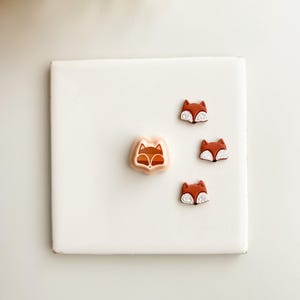 May include: A collection of fox-shaped embellishments in shades of brown, white, and tan. The fox heads are arranged on a white square surface. The image includes a larger fox-shaped item and three smaller ones.