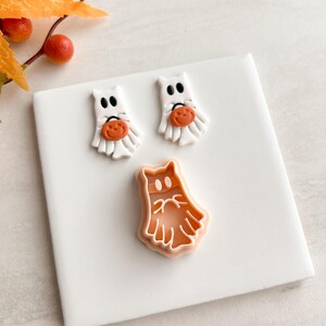 Könnte beinhalten: Drei Halloween-Dekorationen auf einer weißen Oberfläche. Zwei weiße Geisterfiguren mit schwarzen Augen und orangefarbenen Kürbisdetails. Ein dritter, größerer Geister-Ausstecher in Orange.