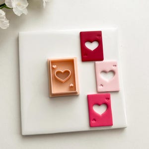 May include: A collection of heart-shaped playing card designs in various shades of pink and red. A peach-colored cookie cutter with a heart-shaped cutout is also present. The cards are arranged on a white surface, suggesting a crafting or baking theme.