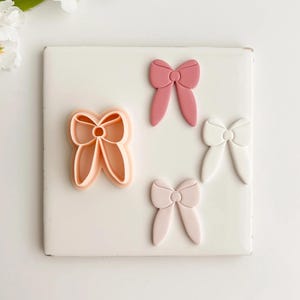 May include: A collection of bow-shaped cookie cutters and fondant decorations in various shades of pink and white. One cutter is peach-colored, and three bows are in different shades of pink and white. The items are arranged on a white square surface.