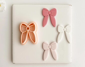May include: A selection of bow-shaped cookie cutters and fondant decorations in shades of pink and white. One cutter is peach-coloured, and three bows are in different shades of pink and white. The items are arranged on a white square surface.