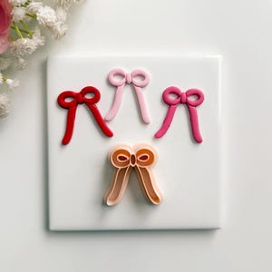 May include: Four bow-shaped cookie cutters in shades of red, pink, and peach are arranged on a white surface. The cutters are made of plastic and are designed for creating bow-shaped cookies or decorations. The bows are in various sizes.