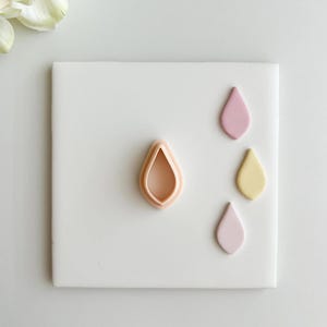 May include: A teardrop-shaped cookie cutter in peach and a few teardrop-shaped cookies in pink and yellow are arranged on a white square tile. The cookie cutter is in the center, with the cookies to the right.