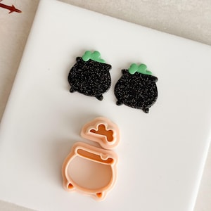 May include: Two black cauldron-shaped earrings with green bubbling potion and a light pink cauldron-shaped cookie cutter. The earrings have a glittery texture. The cookie cutter is in two pieces.