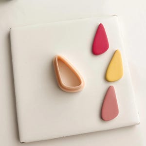 May include: A light peach-colored polymer clay cutter next to three teardrop-shaped clay pieces in shades of red, yellow, and pink. The items are arranged on a white surface.