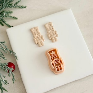 May include: Three tan-colored nutcracker cookie cutters. Two are the nutcracker shape, and one is the cutter. The cutters are on a white surface with a sprig of greenery and a red ornament in the background. The nutcrackers have a crown and a mustache.