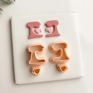 May include: Four pink and peach-colored cookie cutters in the shape of stand mixers. Two are solid pink, and two are peach-colored outlines. The cutters are arranged on a white surface.