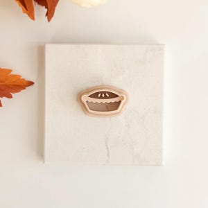 May include: A light brown pie-shaped cookie cutter with a fluted edge and three small dots. The cutter is on a white surface, with a pumpkin and autumn leaves in the background.