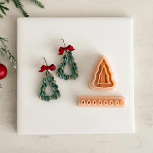 Könnte beinhalten: Zwei grüne und goldene Weihnachtsbaum-Ohrringe mit roten Schleifen, ein Ausstecher in Baumform und ein kleines rechteckiges Werkzeug mit acht Löchern. Die Ohrringe bestehen aus kleinen, geschichteten grünen Teilen mit goldenen Akzenten.
