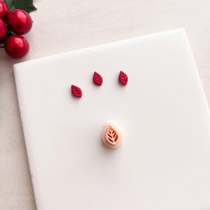 Puede incluir: Una baldosa cuadrada blanca con tres pequeños objetos rojos en forma de hoja y un cortador en forma de hoja. Las bayas y hojas de acebo están en la esquina superior izquierda. Las formas de las hojas tienen vetas detalladas.