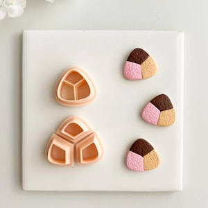 May include: A set of three-part cookie cutters and three finished clay pieces. The cutters are peach-colored plastic, and the clay pieces are brown, pink, and tan, resembling a triangular shape. The items are on a white surface.
