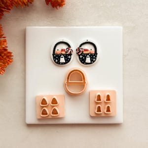 Peut inclure: Deux boucles d'oreilles ovales blanches avec un motif de panier noir rempli d'un fantôme, d'une citrouille, de bonbons maïs et d'une sucette. Trois emporte-pièces en plastique pêche sont inclus : un grand panier, un bloc rectangulaire avec quatre formes de bonbons maïs et un bloc rectangulaire avec quatre formes de fantômes.