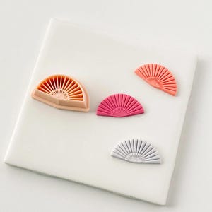 May include: Four fan-shaped objects in peach, pink, and grey are displayed on a white surface. The fans have a ribbed design, with colours ranging from light to dark.