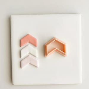 May include: A collection of chevron-shaped items in shades of pink and white, arranged on a white square. The items include three solid chevrons and a chevron-shaped cookie cutter, all displayed against a clean, bright background.