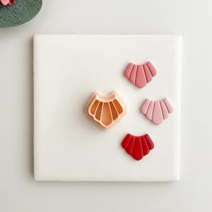 May include: A collection of seashell-shaped cookie cutters and cookies in shades of pink and red. The cookie cutter is a light orange color. The cookies are in various shades of pink and red. The items are arranged on a white surface.