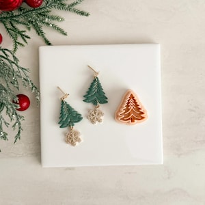 May include: Green Christmas tree earrings with gold snowflake charms, alongside a tree-shaped cookie cutter. The earrings and cutter are presented on a white square, with festive greenery and red baubles in the background, evoking a holiday theme.