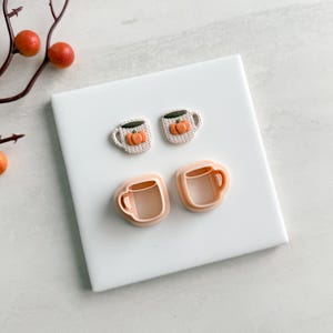 以下が含まれることがあります： 秋をテーマにしたマグカップのデザイン2セット。 上段には、オレンジ色のカボチャのデザインが施された2つの白いマグカップがあります。 下段には、2つの明るいオレンジ色のマグカップ型のクッキーカッターが表示されています。 アイテムは、オレンジ色のベリーの小枝が付いた白い表面にあります。