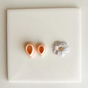 May include: A light peach-colored petal-shaped cookie cutter set next to a small, multi-layered flower with gray and white petals and a gold center. The items are on a white surface.