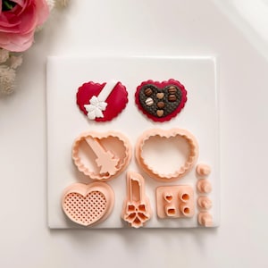 Op de afbeelding: Een verzameling roze hartvormige koekjessnijders en twee hartvormige fondantdecoraties. Eén decoratie is rood met een witte strik, de andere rood met bruine chocoladesnoepjes. De uitstekers zijn in verschillende vormen en maten.