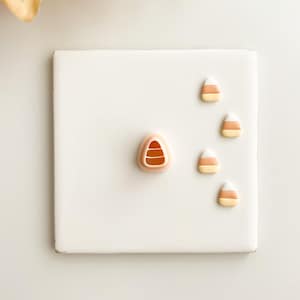 Puede incluir: Una baldosa cuadrada blanca con cuatro decoraciones de caramelos de maíz. Un caramelo de maíz es más grande y tiene un diseño detallado. Los otros tres son más pequeños y sencillos. Las decoraciones de caramelos de maíz son de color naranja, amarillo y blanco.