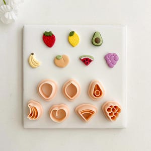 May include: A collection of fruit-shaped cookie cutters, including a strawberry, lemon, avocado, banana, peach, watermelon, and grapes. Also shown are various geometric shapes. The cutters are in shades of red, yellow, green, and peach.