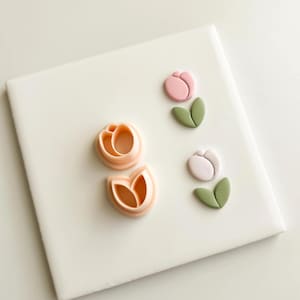 Puede incluir: Un conjunto de cortadores de galletas y moldes de fondant con forma de tulipán en varias etapas de finalización. Los cortadores son de color melocotón, mientras que los tulipanes terminados son rosas y blancos con hojas verdes, dispuestos sobre una superficie blanca.