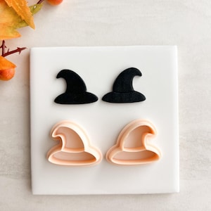 May include: Four witch hat cookie cutters. Two are black, and two are light peach. The cutters are arranged on a white square tile, with orange leaves and berries in the background. Perfect for Halloween baking.
