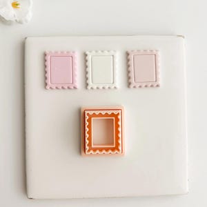 Puede incluir: Cuatro cortadores de galletas con forma de sello en tonos rosa, blanco y naranja. Los cortadores tienen un diseño de borde festoneado. Los cortadores están dispuestos sobre una superficie blanca, con algunas flores blancas en el fondo.
