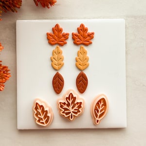 May include: A set of six leaf-shaped clay pieces in shades of orange, yellow, and brown, arranged in pairs. Below them are three light peach-colored plastic cutters shaped like a jagged leaf, a maple leaf, and an oval leaf with vein details.