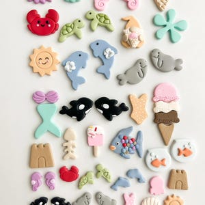 Puede incluir: Una colección de adornos en miniatura coloridos con temática oceánica. El surtido incluye criaturas marinas como caballitos de mar, delfines y cangrejos, junto con conos de helado, castillos de arena y una cola de sirena. Los colores son pastel y brillantes.