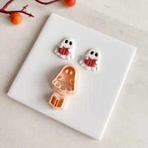 May include: A Halloween-themed image featuring three small, white ghost figures with red books, and two cookie cutters. The cookie cutters are in the shape of a ghost and a book. The items are on a white surface.