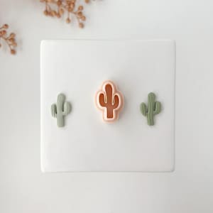 Puede incluir: Tres objetos con forma de cactus en tonos verdes y melocotón están dispuestos sobre una superficie blanca. El objeto central es un cortador de galletas con forma de cactus de color melocotón. Dos objetos verdes con forma de cactus están a cada lado.