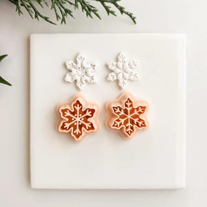 May include: Four snowflake-shaped cookie cutters. Two are white, and two are light orange with a brown snowflake design. The cutters are arranged on a white square surface, with green and red Christmas decor in the background.