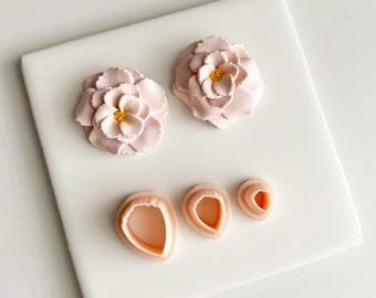 May include: A selection of light pink flower cake decorations and petal cutters. Two large, fully bloomed flowers with yellow centres are displayed with three petal cutters in different sizes. The items are arranged on a white surface.