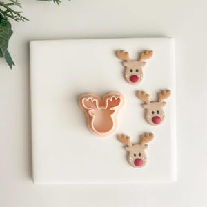 May include: A light pink reindeer cookie cutter next to three reindeer cookies with brown antlers and red noses. The cookies are on a white square surface, with a sprig of greenery in the upper left corner.