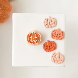 May include: A collection of five pumpkin-shaped decorations in varying shades of orange and peach. Each pumpkin features a carved jack-o'-lantern face. The pumpkins are arranged on a white surface, perfect for Halloween decor.