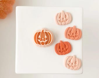 May include: A collection of five pumpkin-shaped decorations in varying shades of orange and peach. Each pumpkin features a carved jack-o'-lantern face. The pumpkins are arranged on a white surface, perfect for Halloween decor.