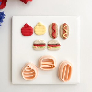 Può includere: Una collezione di formine per biscotti e prodotti da forno a tema cibo. Il set comprende contenitori di condimenti rossi e gialli, hot dog con condimenti rossi e gialli e hamburger. Ci sono anche tre forme di formine per biscotti non cotti.