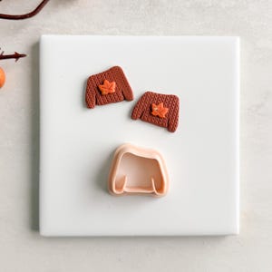 May include: A collection of fall-themed polymer clay creations. Two miniature brown sweaters with orange leaf accents sit above a sweater-shaped cookie cutter. The items are arranged on a white square surface.
