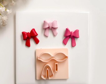 May include: A top-down view shows three decorative bows in red, pink, and fuchsia. A light peach-coloured cookie cutter with bow shapes is also present. The items are arranged on a white square surface.